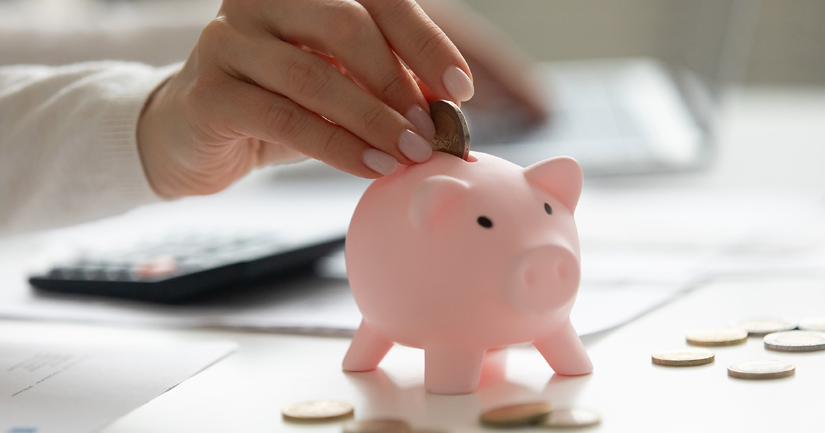 Close up of a woman holding a smartphone and putting a coin in a pink piggy bank. The image prompts the question what is financial aid?