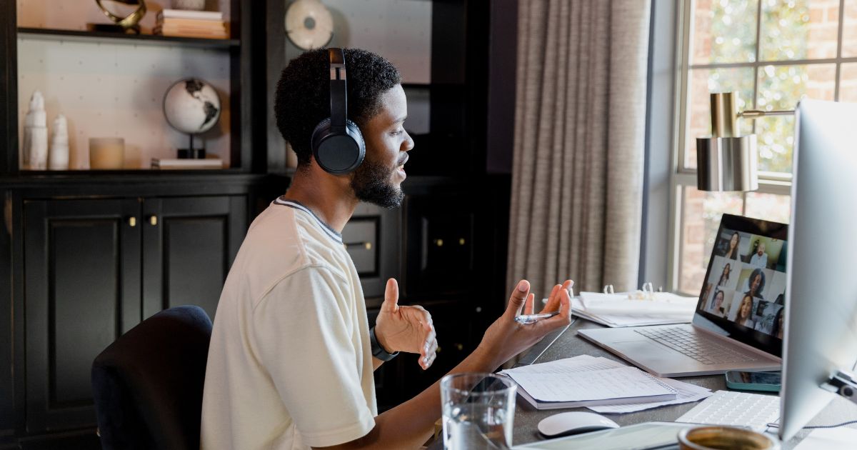 A Black man wearing headphones sitting at a desk and gesticulating during a live virtual meeting by a window. He is embodying successful virtual meeting tips.