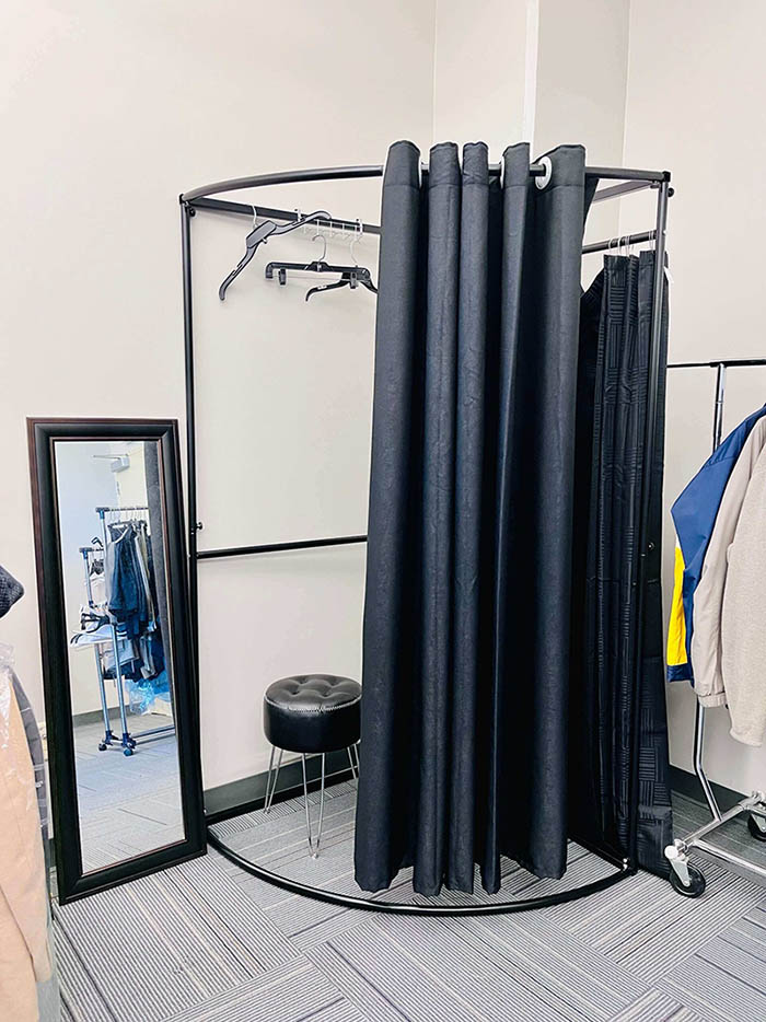 A dressing room with a curtain and a stool inside it tucked in a corner next to a clothes rack and a mirror.