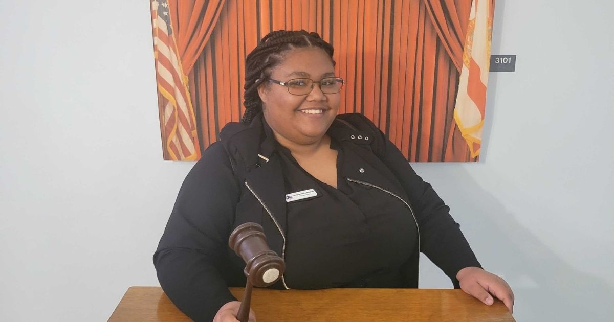 A smiling Black woman, Annjeannette Mitchell, wearing glasses and black sweatshirt with a name tag, stands behind a podium holding a gavel and displaying her SGA leadership strategies.