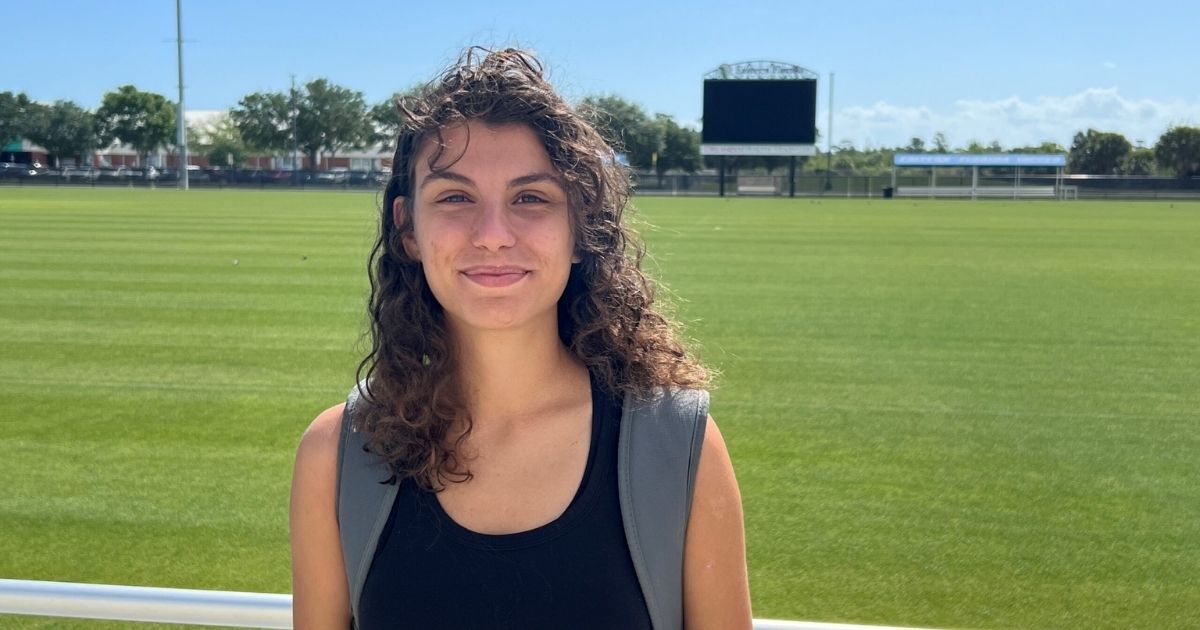 Akatarinha Papagiorgio, a woman with long curly hair wearing a black tank top and a backpack, stands in front of a grassy athletics field for her athlete spotlight.