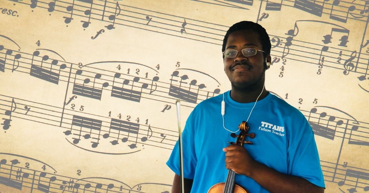 Campus violinist Anthony Hires, a Black man wearing glasses and a Titans Future Teacher shirt, stands in front of sheet music holding his violin.