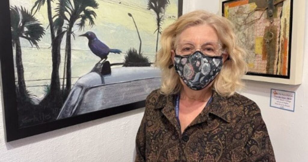 EFSC art instructor Georgi Gelalles, a woman with shoulder-length blonde hair, glasses, a face mask, and a floral blouse, stands beside a painting of a bird.