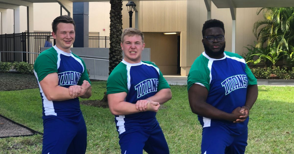The three male members of the EFSC Cheer Team stand in a row flexing their biceps with various expressions conveying comedy and seriousness. They are wearing blue-green-white athletic uniforms that say "titans" on the front.
