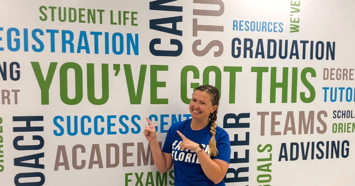 Jaime Braudrick, a smiling woman with blonde hair in a braid, hoop earrings, and an Eastern Florida shirt, points with both hands to a word cloud on a wall, giving first-term at EFSC advice.