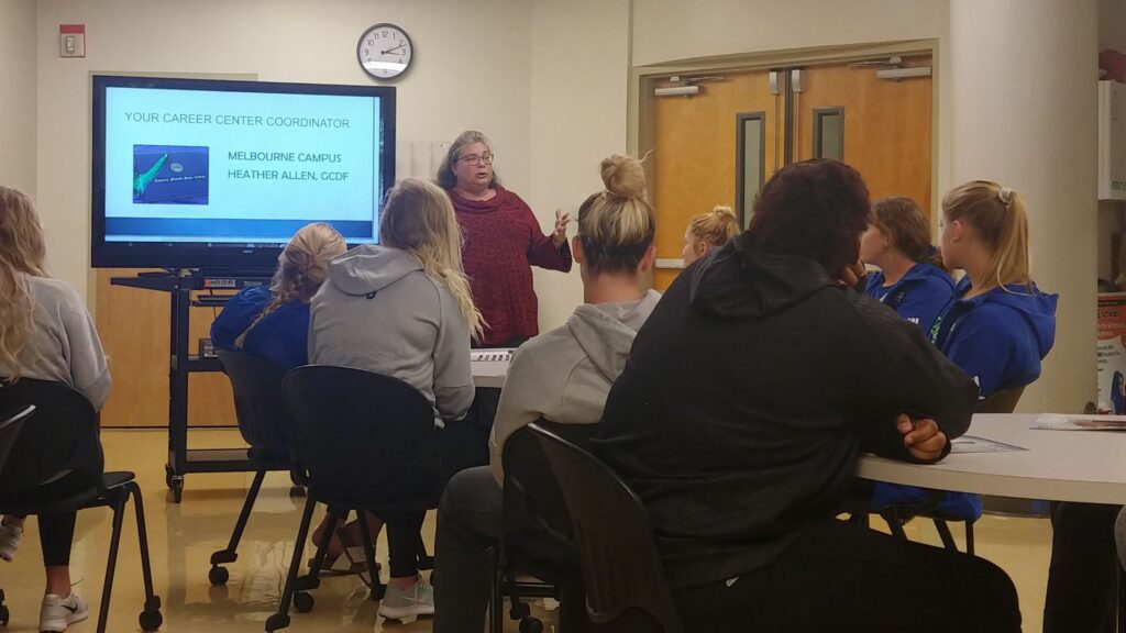 A white woman in glasses and a red sweater standing next to a TV screen that reads "Your Career Center Coordinator Melbourne Campus Heather Allen, GCDF" and presenting to a room full of students.