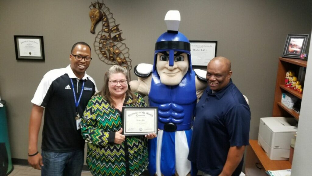 Two Black men in polo shirts (Ancel Robinson, Jr. and Myron Campbell) standing next to a white woman in glasses and a chevron-patterned blouse (Heather Allen, who is holding up her Recruiter of the Month certificate) and Mr. Titan.