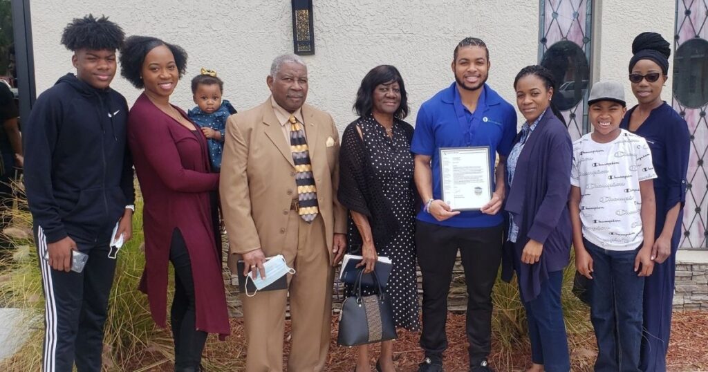 A Black man in a blue polo shirt, Xavier Edwards, holds up a framed certificate documenting his success story. He is surrounded by eight people: his friends and family.