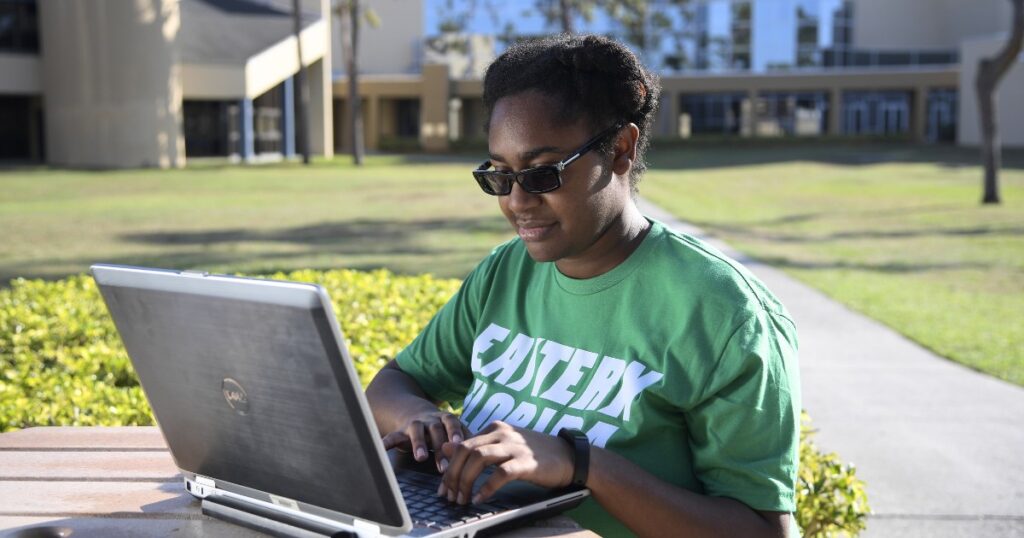 A young woman wearing a green EFSC shirt sits at an outdoor table with a laptop open, writing a professional email