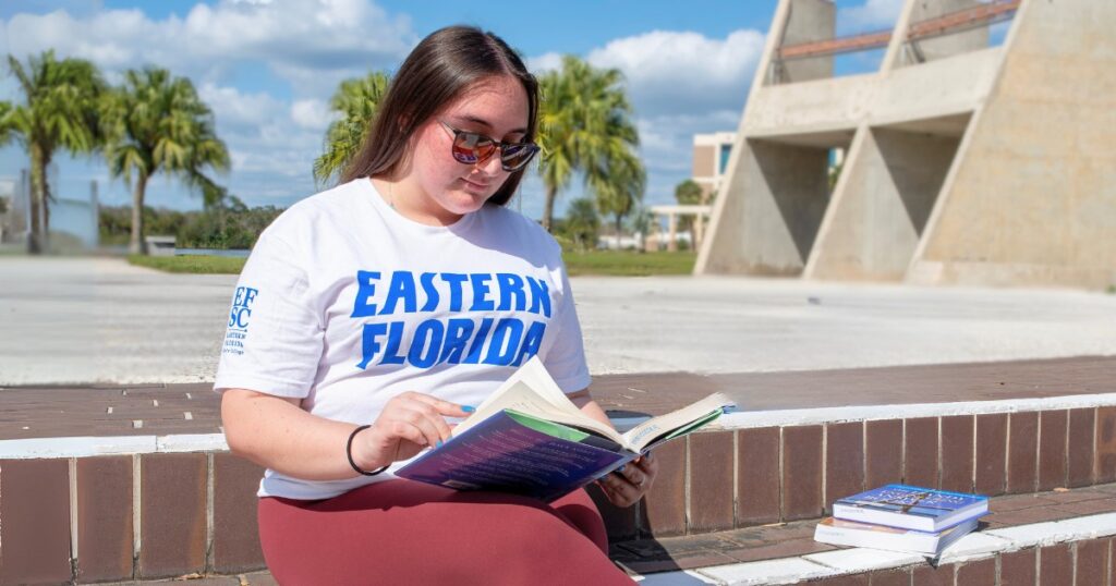 A female student with long brown hair and sunglasses wearing an EFSC shirt reads from a textbook while sitting on stone stairs