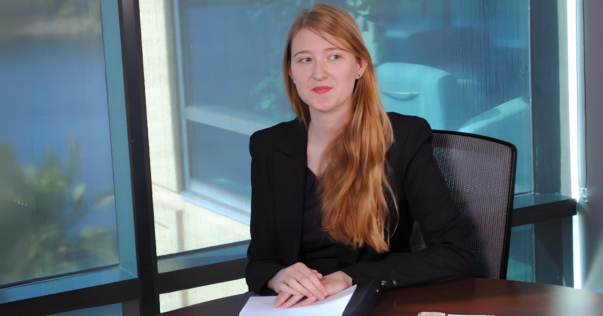 A young woman in a blazer sitting with hands folded over a stack of documents on a polished desk. She has long red hair flourishing over her shoulder.