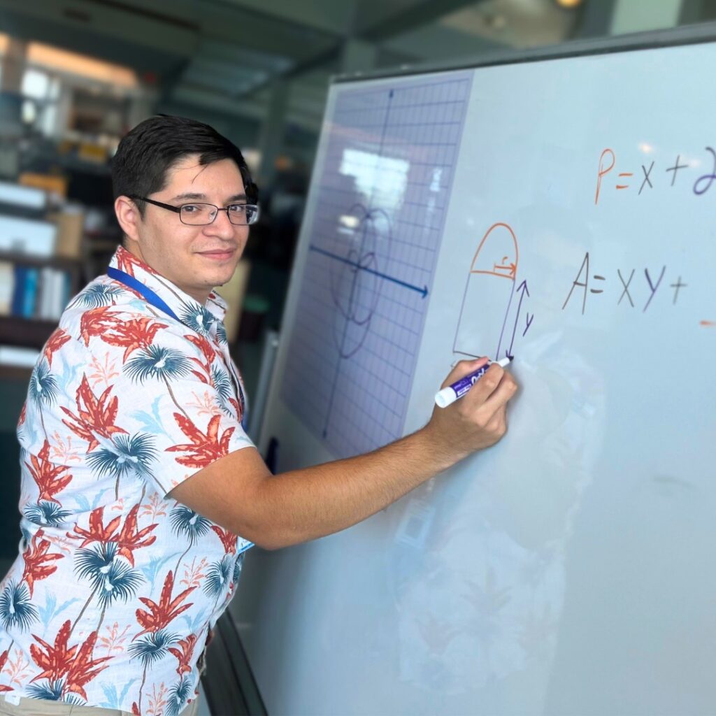 Daniel Garcia, a young man wearing a Hawaiian printed dress shirt and glasses, writing a math equation on a white board.