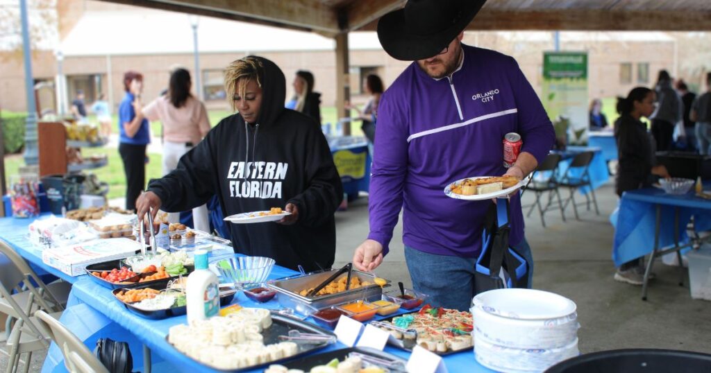 Two students wearing Eastern Florida State College clothing using tongs to select food off of an outdoor table, including wraps.