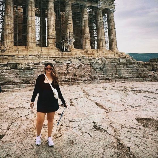 Hailey, a student, stands in front of Greek ruins on a cloudy day