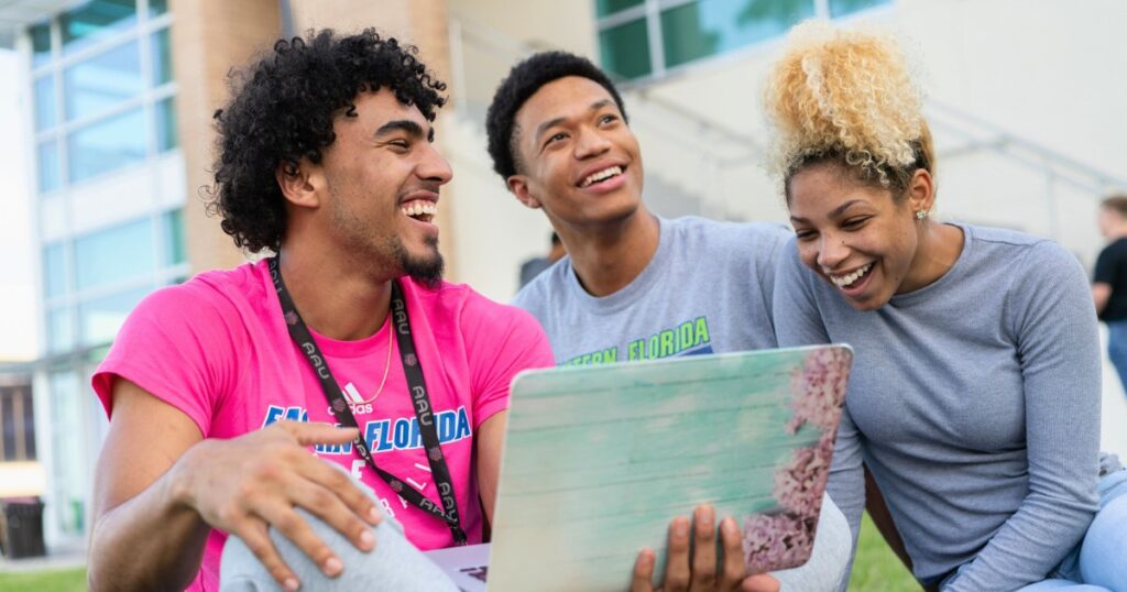 Three students laughing and smiling around a laptop while sitting outside of the Student Union building on the lawn.