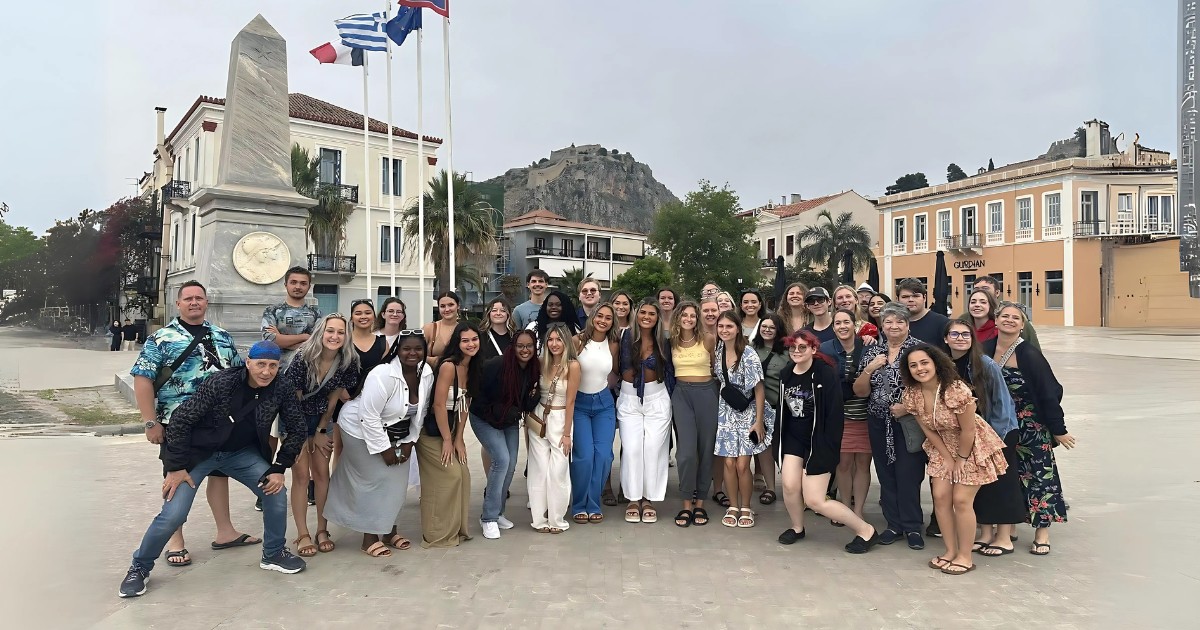 A group of diverse students standing in an open square in Greece with a mountain and cultural architecture and flags in the background.