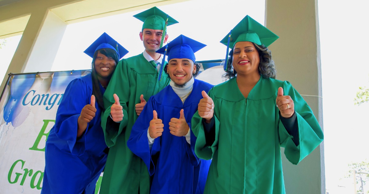 A group of four students of different ages and ethnicities dressed in green and blue gowns smile and give a thumbs up at the camera