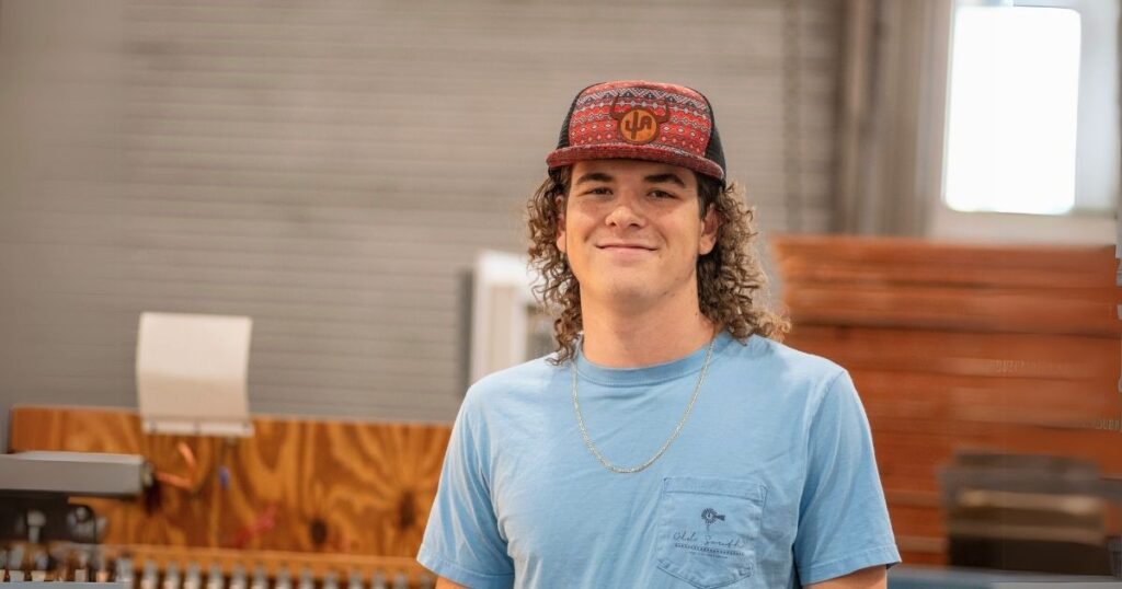 A young man in a light blue t-shirt with long curly hair and a red ballcap