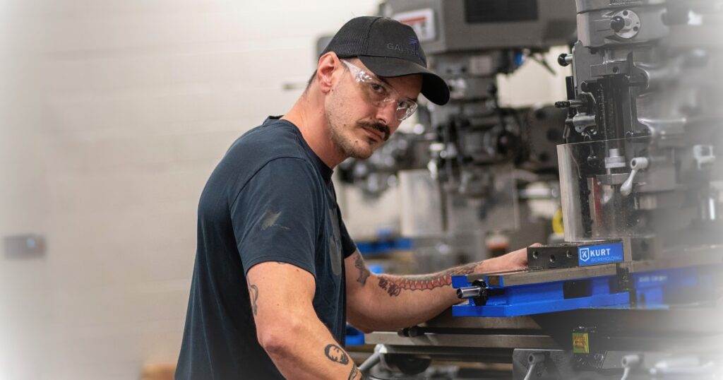 A male student wearing a ballcap and turned toward the camera while working on a CNC machine apparatus