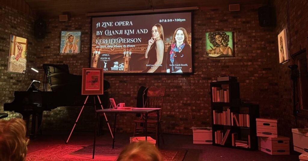 A small stage designed to look like a woman's study, with artistic portraits of women on the walls, a piano in the corner, and books on shelves; a table sits central with a projector in the back with information about the A Zine Opera. The stage lighting is red.