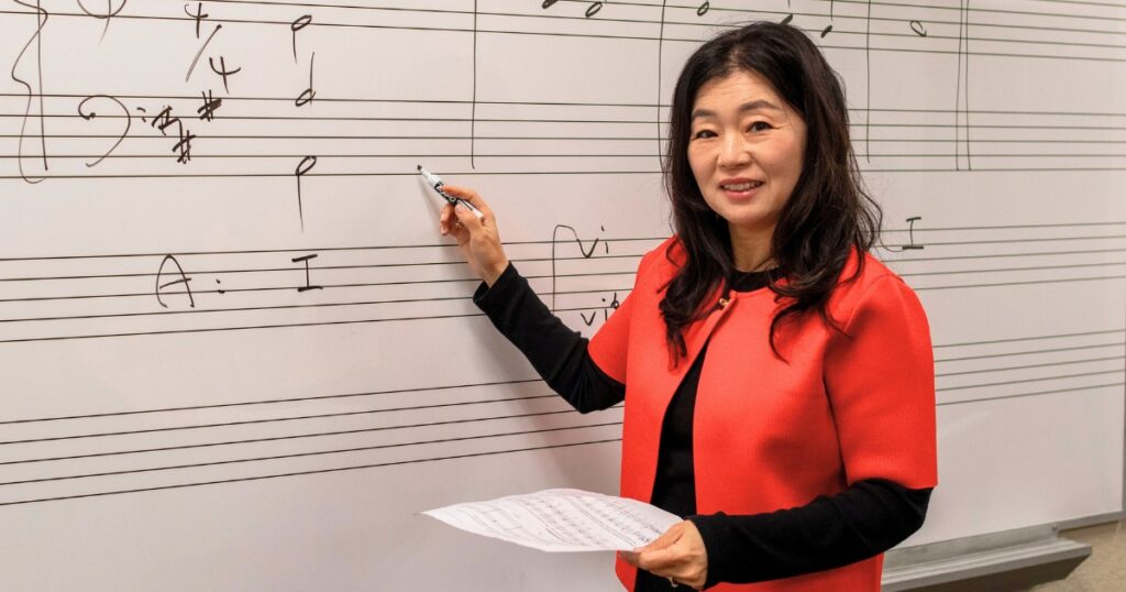 Professor ChanJi Kim, a woman with shoulder-length black hair and a red jacket, holds a piece of sheet music and writes musical notes on a staff board