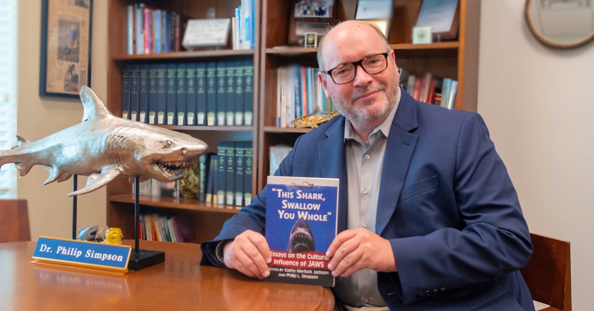 Dr. Philip Simpson is a bald man wearing glasses and a navy blue jacket holding a copy of his Jaws anthology while posing next to a silver statue of a shark with a placard of his name
