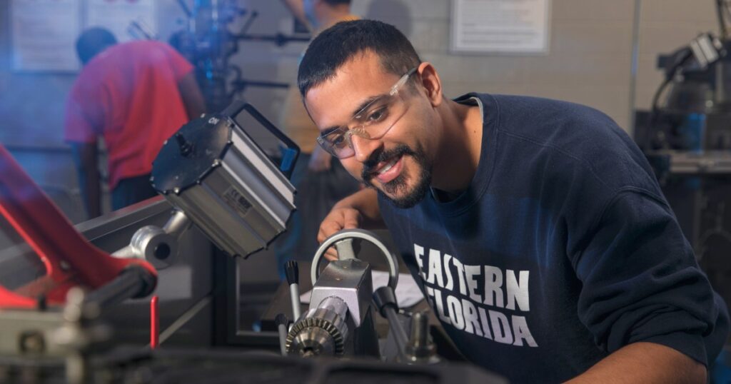 A man with short-cropped hair and a mustache and beard bends over a piece of machinery on a table, working on CNC equipment.