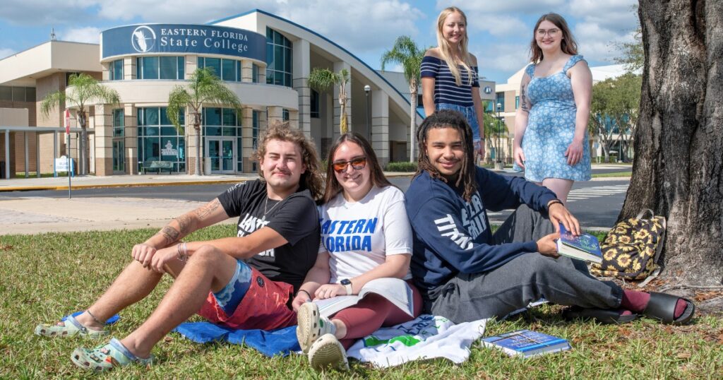 A group of diverse students sit on the grass and stand in the background, which shows an EFSC building