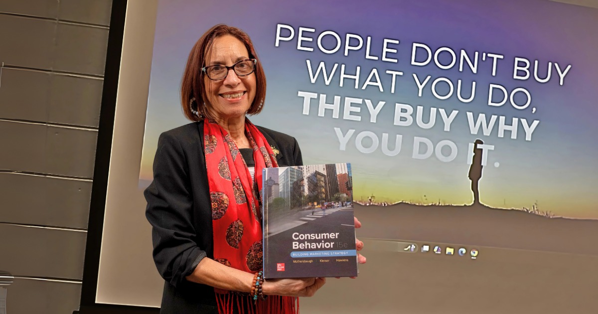 Dr. Nicole DeCaro, a woman with jaw-length straight brown hair and glasses, wearing a professional black blazer and red scarf while holding a business textbook