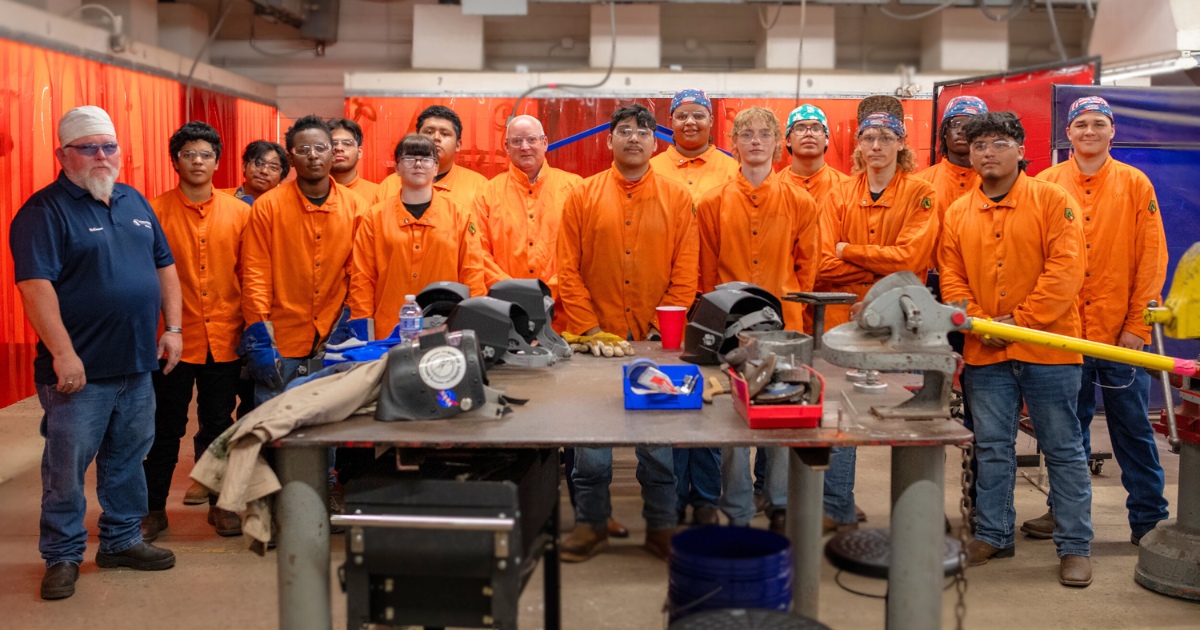 A group of diverse students wearing orange welding uniforms standing with their teachers behind a welding bench
