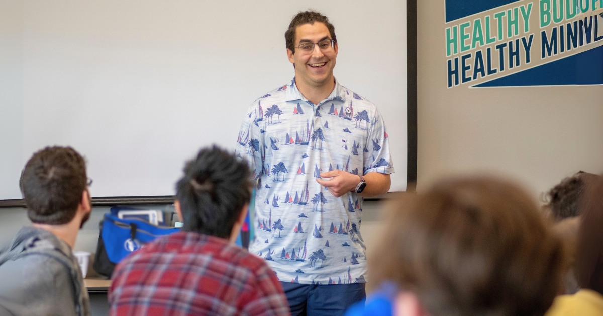 A group of students in a classroom listen to speaker, Daniel Perkins, a young man with a white pattered shirt with a black ponytail.