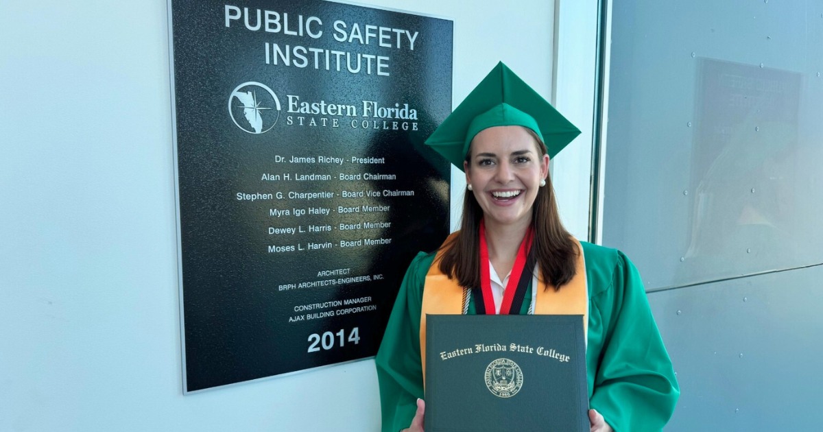Kaylin Mitchell, a woman wearing a green graduation uniform and cap with long straight brown hair and holding her commencement diploma case in front of a plaque of EFSC's Public Safety Institute