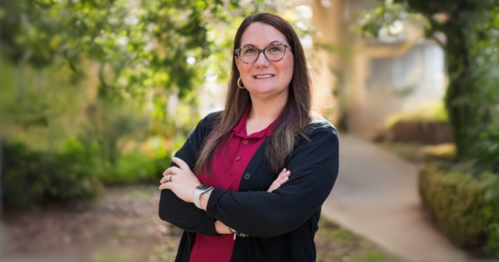 Misty Shirah, a woman with long straight brown hair, wearing a red shirt and black blazer