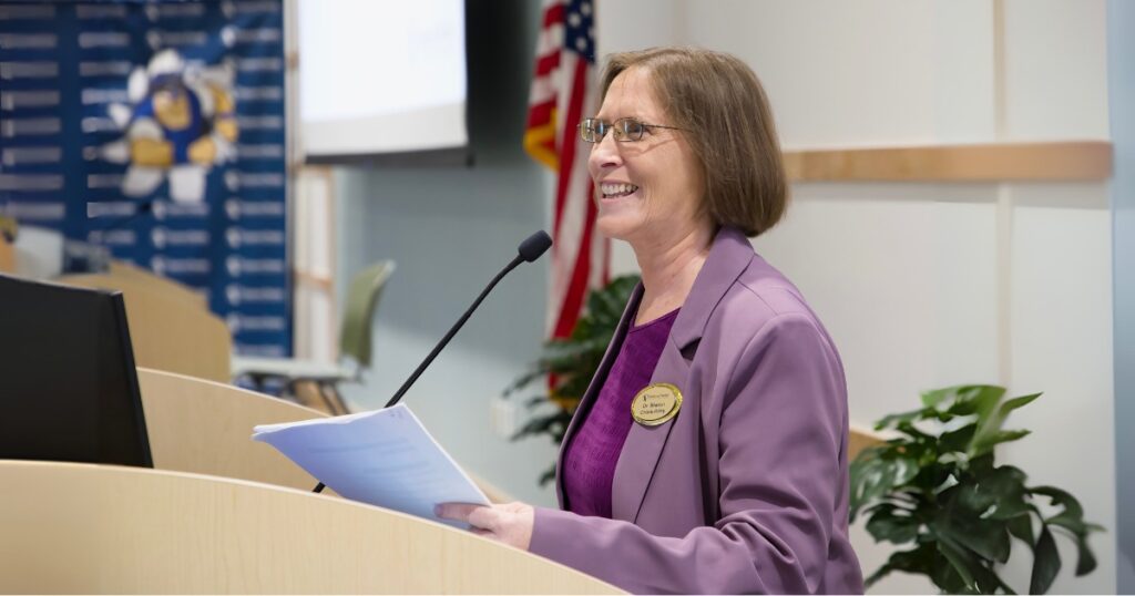 An image of Sharon Cronk-Raby wearing a purple blazer standing at a podium and giving a presentation while holding a collection of papers in her hand. She smiles while speaking into a microphone.