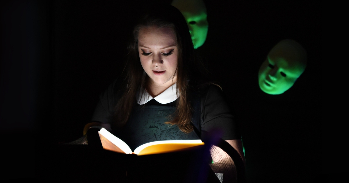 A female student with long straight brown hair reads from a book on a podium with low lighting. Green masks hover behind her ominously.