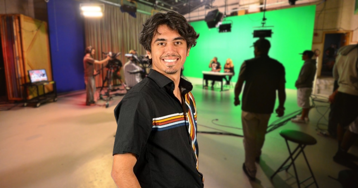 Enrique J. Molinares stands in the foreground of a television production studio, wearing a black short-sleeve shirt with multicolored stripe accents. Behind him, camera operators and crew members work on a set featuring a large green screen, studio lighting, and two people seated at a desk, illustrating a live media production environment.