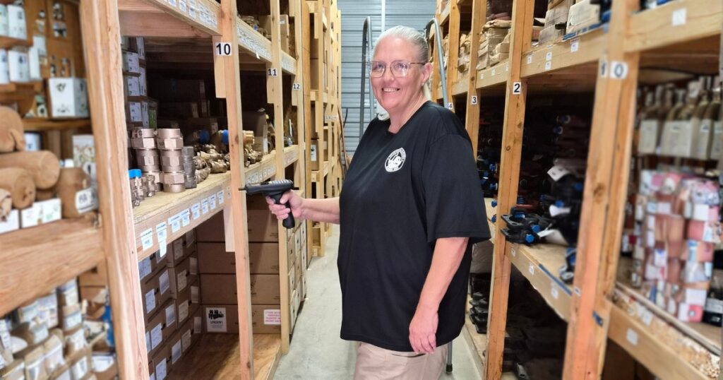 Vicki Perry, a woman wearing a black polo and safety goggles standing in a warehouse taking inventory