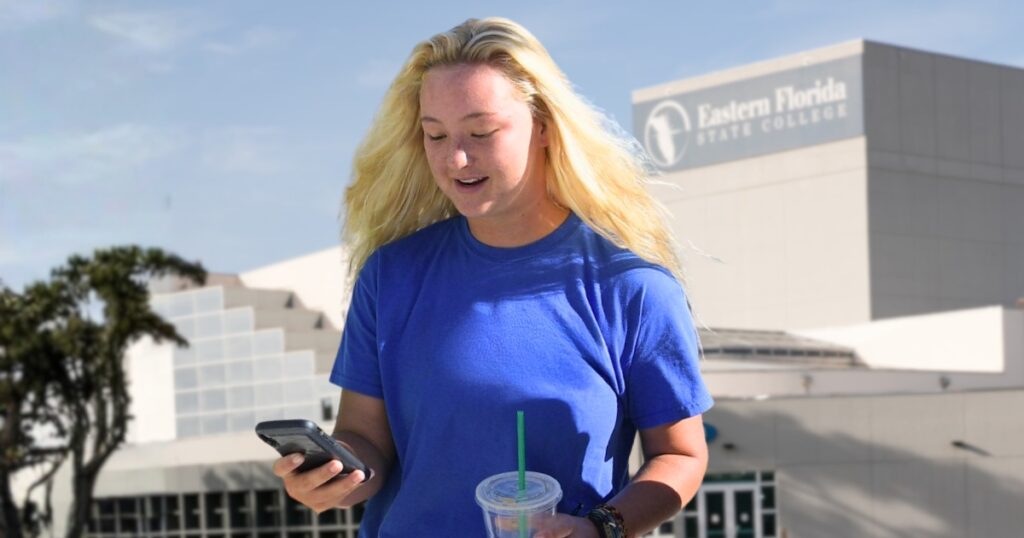 A blond student with a blue shirt texts on her phone while walking across EFSC Melbourne campus
