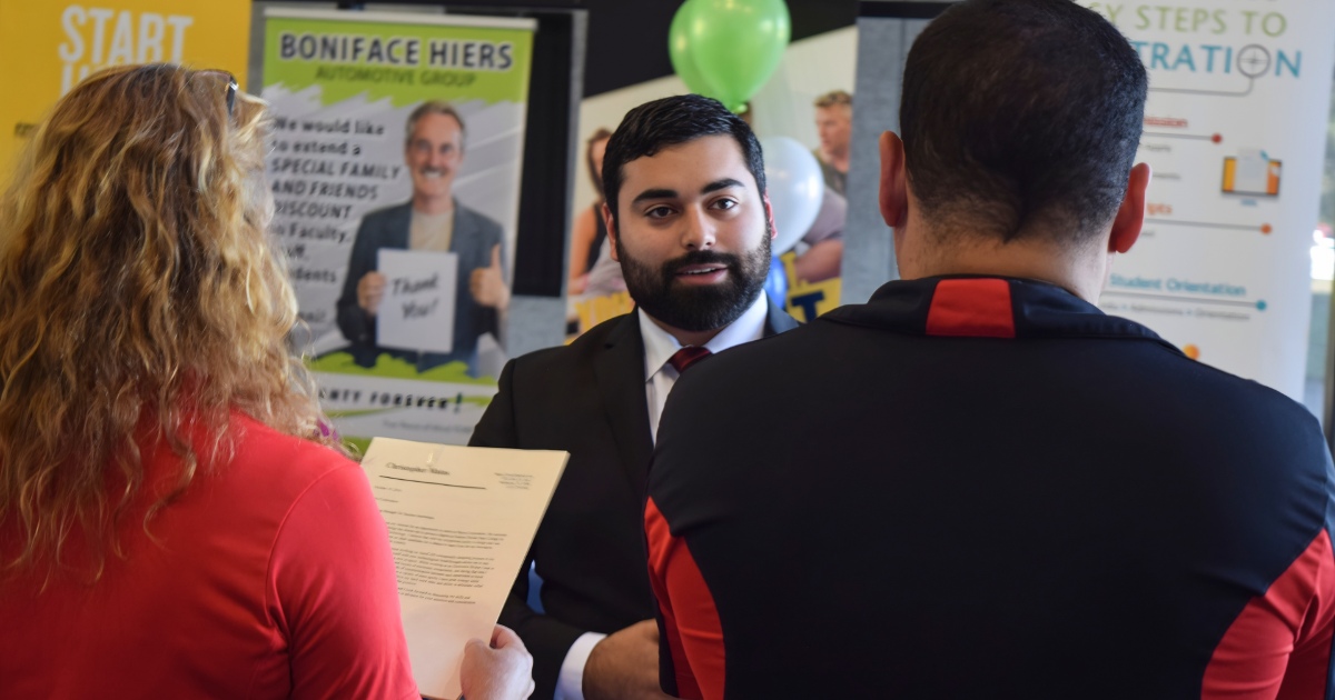 A bearded college student in a business suit speaks to two recruiters at a job fair as they review his resume.