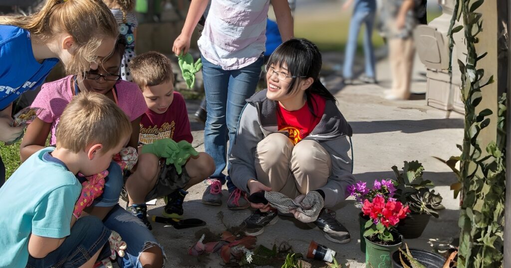 A college student wearing a Rotaract shirt kneels with grade-school students showing them how to pot plants