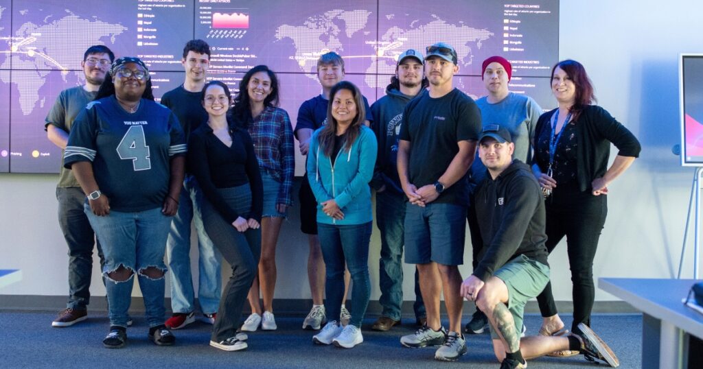 A group of cybersecurity college students standing in front of a cybersecurity TV