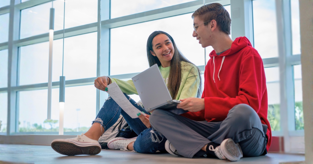 Two busy college students sit on a polished floor in a bright building with large windows, working together with a laptop and a sheet of paper while wearing casual clothing.