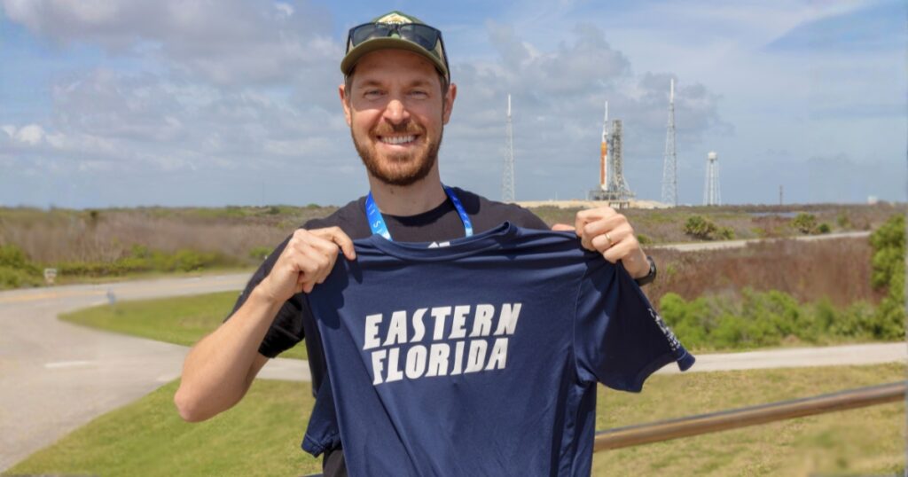 Wade Dauberman, an EFSC instructor wearing a ballcap and lanyard, holding a navy blue t-shirt with the EFSC logo, standing outside with the shuttle launch pad in the background