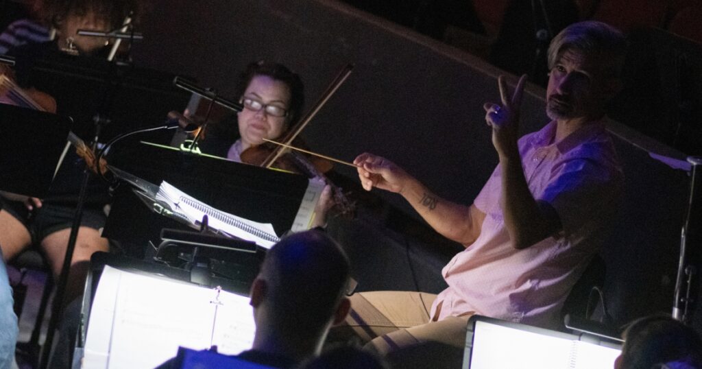 Jon Oxford leads musicians from the orchestra pit during a theatrical performance, illuminated by dim stage lighting and music stand lights. A violinist and other orchestra members are visible in the background with sheet music and instruments.