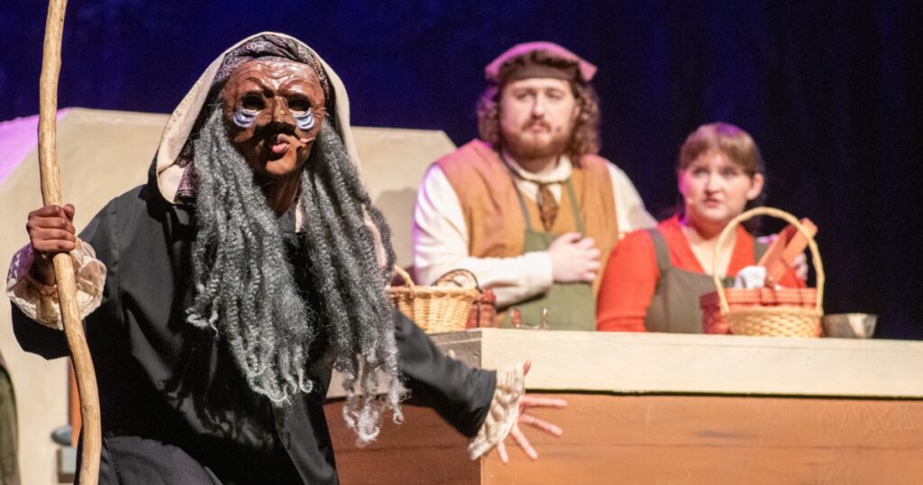 Masked performer holding a wooden staff stands onstage during a theatrical performance of “Into the Woods,” while two costumed performers with baskets watch from behind a market-style set. The stage is lit with warm theatrical lighting against a dark backdrop.
