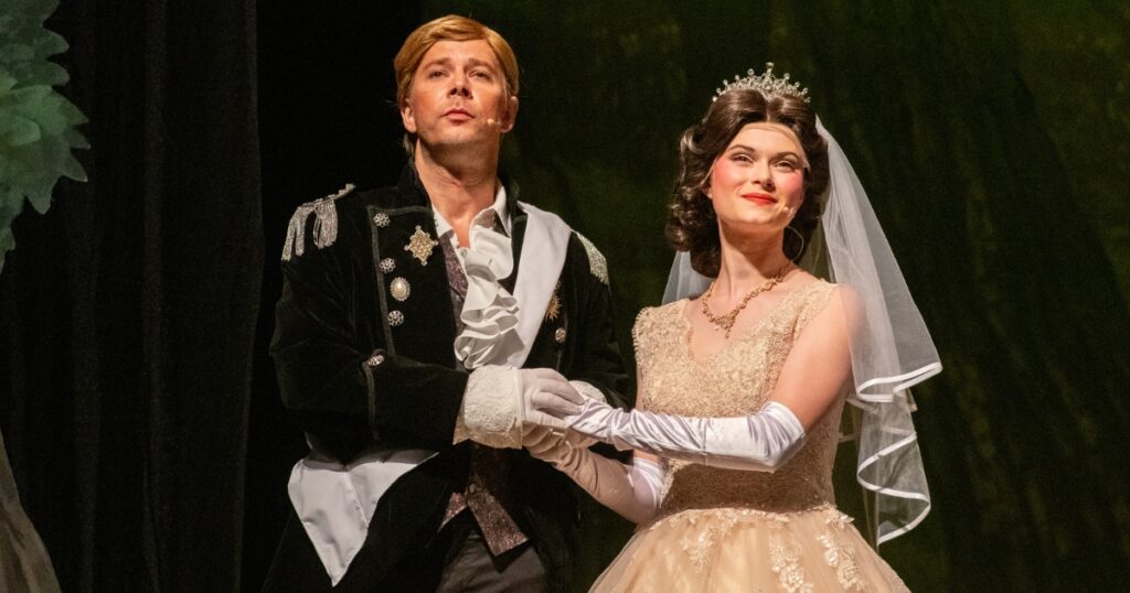 Two performers in formal fairy tale-inspired costumes stand arm in arm onstage during a production of “Into the Woods.” One performer wears a black embellished jacket with white gloves, while the other wears a cream-colored gown with long gloves, a tiara, and a bridal veil. The stage is lit with warm theatrical lighting against a dark forest backdrop.