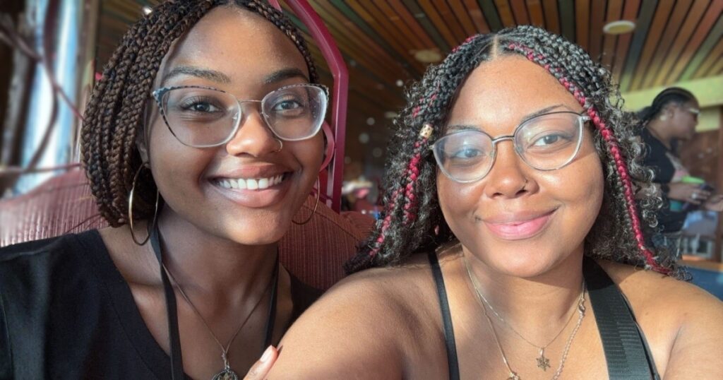 Alayna and Alcenta wearing glasses pose for a close-up selfie while seated indoors at a restaurant or café with wood-paneled ceilings and large windows.