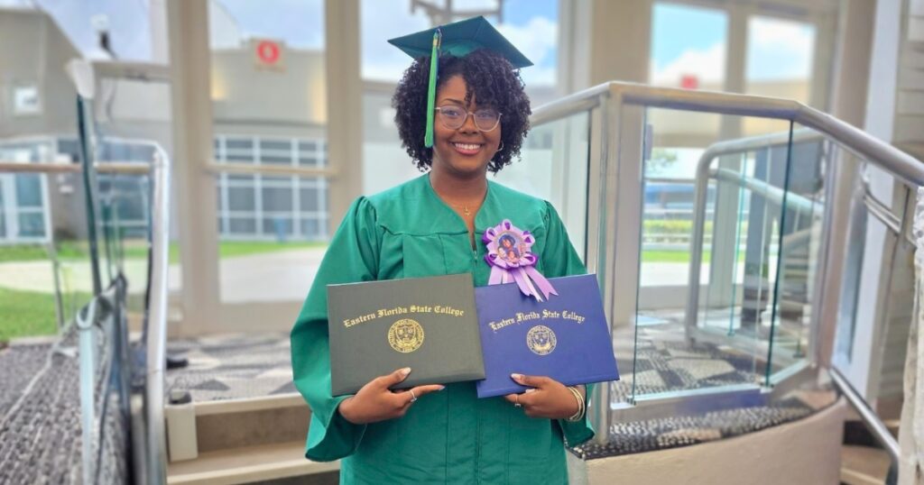 Alayna Castro King in a green graduation cap and gown stands indoors holding two Eastern Florida State College diploma covers near a staircase with glass railings. A purple commemorative ribbon is pinned to the gown.