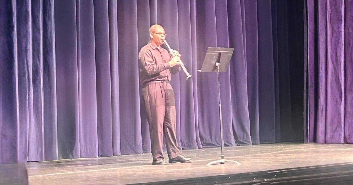 Drawin Flores performing a clarinet solo on a stage, standing beside a music stand in front of purple theater curtains.