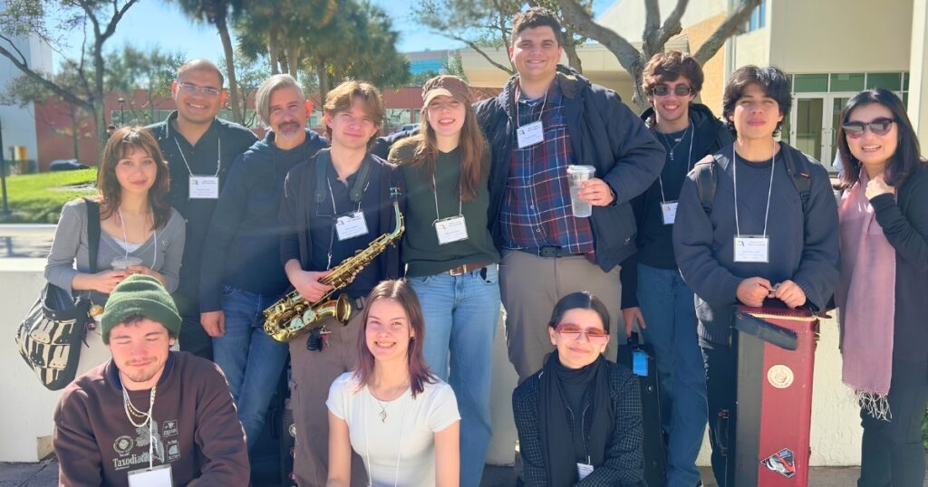 Group of college students wearing name badges gathered outdoors on campus, posing together on steps in sunny weather, with instruments—including a saxophone—and a music case visible.